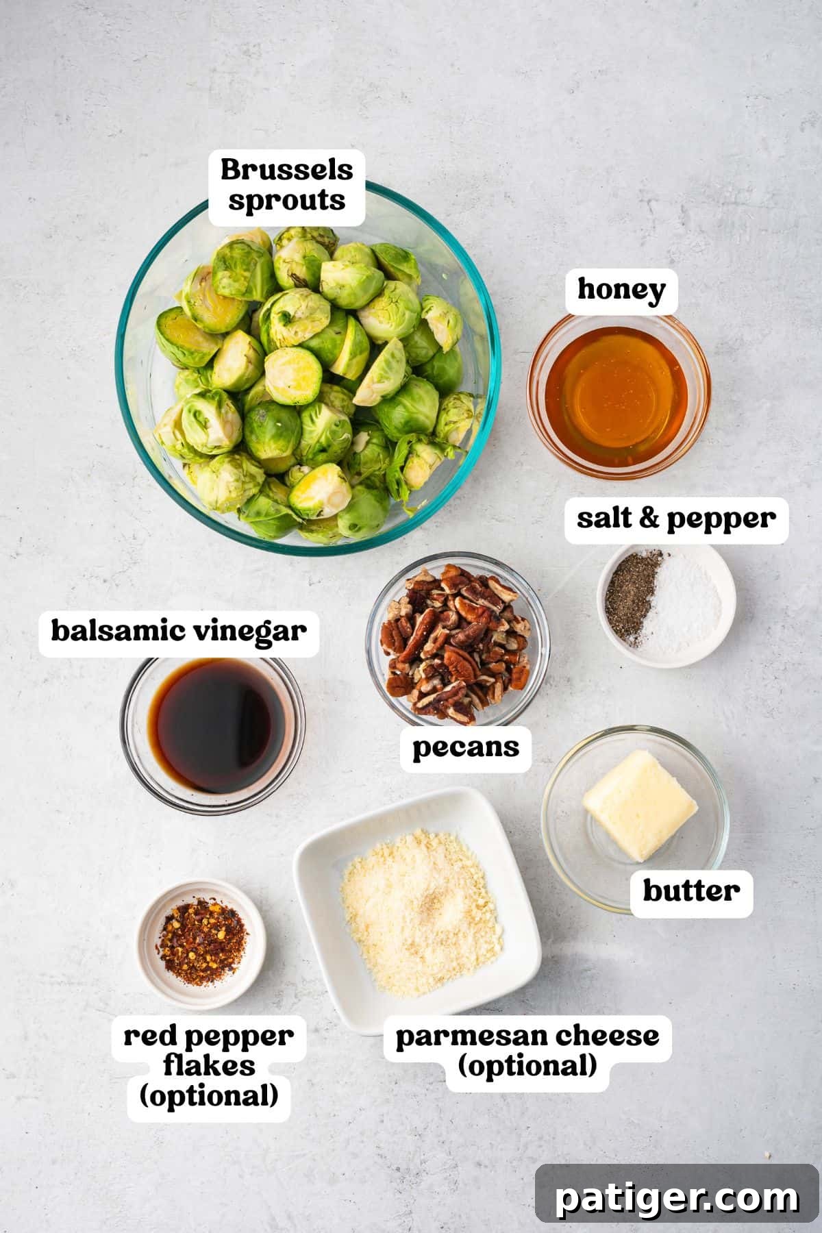 Overhead image of all ingredients labeled: halved Brussels sprouts in a glass bowl, honey, salt and pepper, balsamic vinegar, chopped pecans, butter, red pepper flakes (optional), and parmesan cheese (optional), all arranged on a light surface.