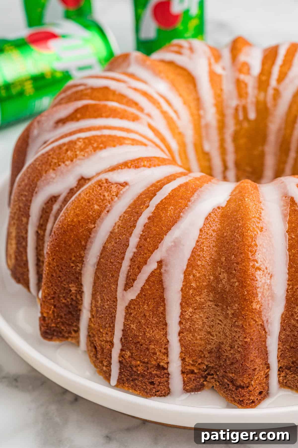A golden brown 7 Up pound cake with white glaze drizzled in thin lines over the top, sitting on a white plate with several green 7 Up cans in the background.
