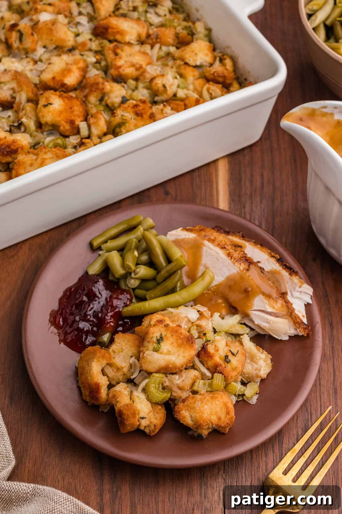 A plate with a serving of golden-brown bread stuffing, slices of turkey breast with gravy, green beans, and cranberry sauce. In the background, a white baking dish of stuffing and a gravy boat are partially visible on a wooden table.