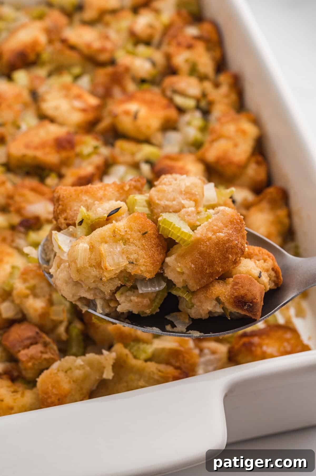 A close-up of a spoonful of homemade stuffing lifted above a white baking dish. The spoon holds rustic toasted bread cubes mixed with chopped celery, onions, and herbs.