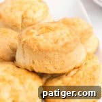 Overhead view of multiple golden brown biscuits piled on a white serving platter, with a honey dipper, bowl of honey, and stacked plates visible in the background.