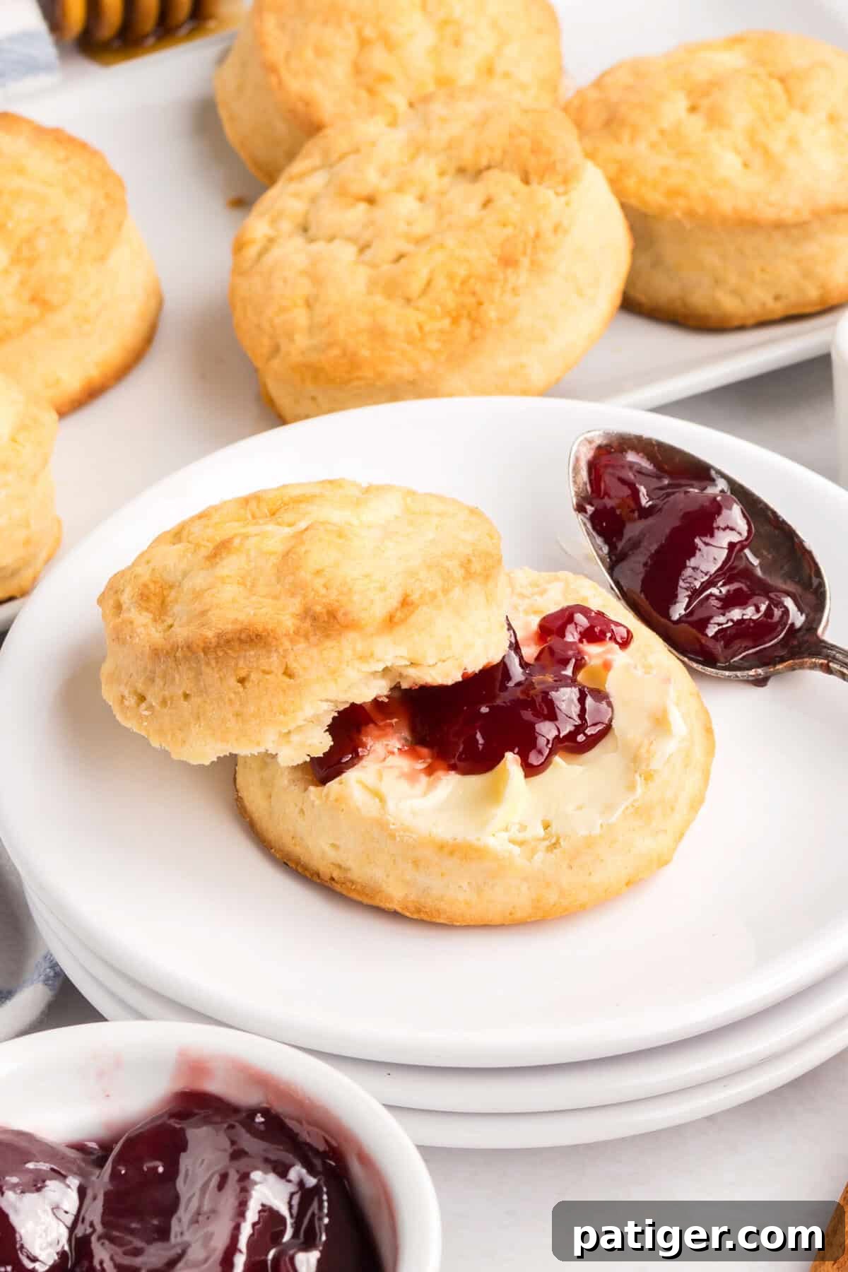 Golden biscuit cut in half and topped with butter and jam, placed on a stack of white plates with more biscuits and jam bowls in the background.