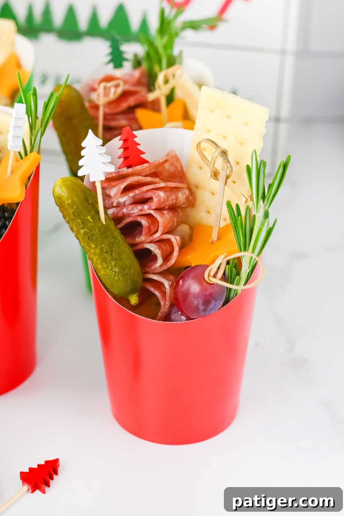 Vibrant Christmas charcuterie cup filled with folded salami skewers, star-shaped cheddar cheese, a gherkin, a fresh grape, a crisp cracker, and a sprig of aromatic rosemary, set against a festive backdrop.