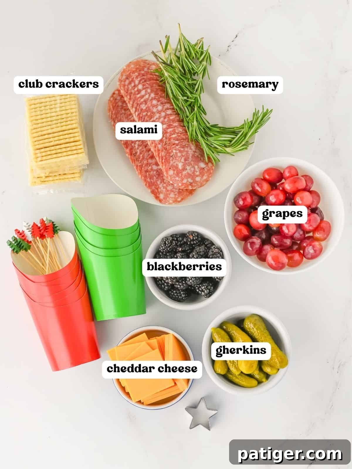 Flat lay image showcasing all labeled ingredients for Christmas charcuterie cups, including Club crackers, fresh rosemary sprigs, neatly folded salami slices, vibrant red grapes, plump blackberries, pre-sliced cheddar cheese, and tangy gherkins. Red and green paper cups with decorative skewers are also visible, ready for assembly.
