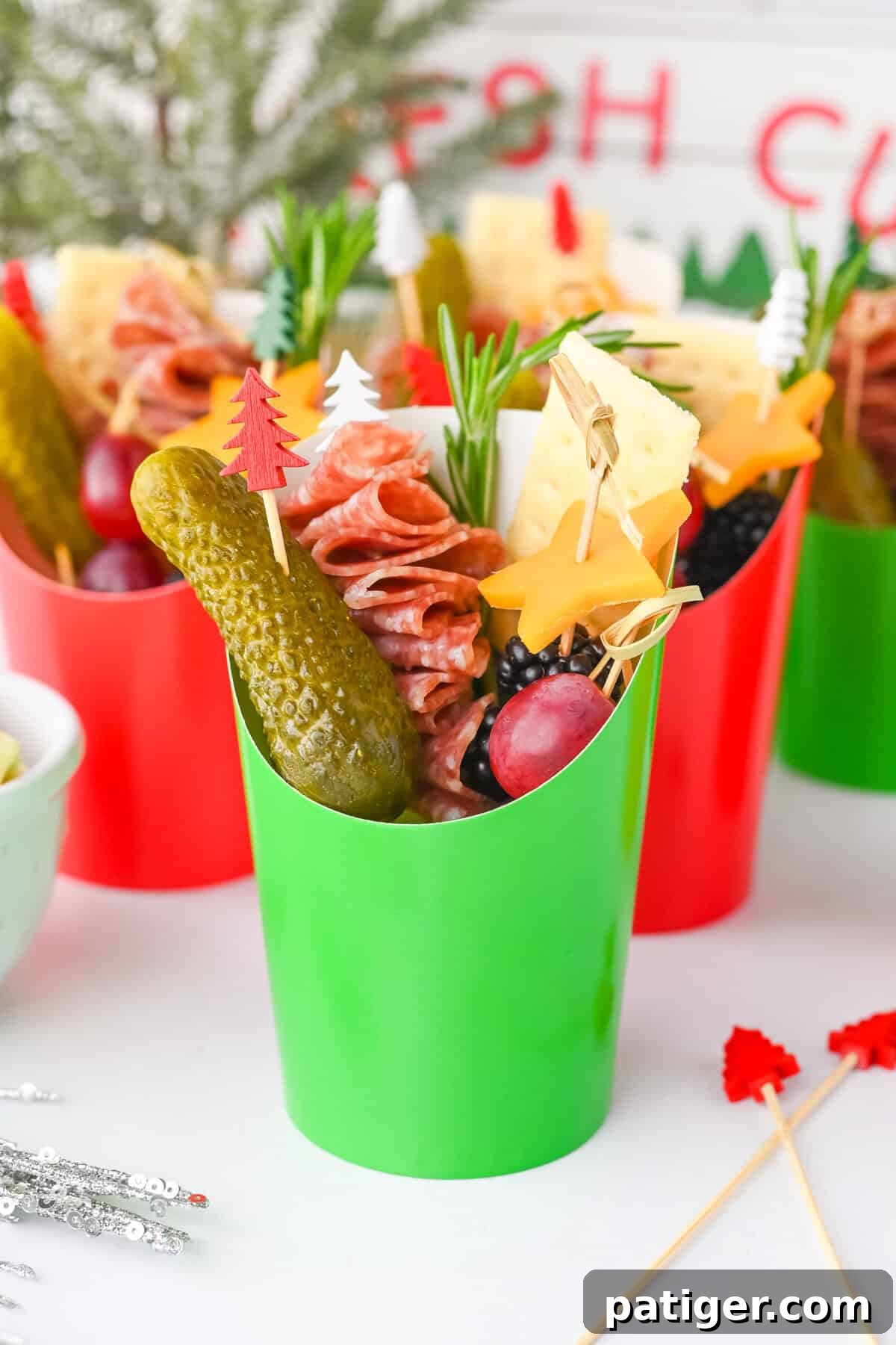 A vibrant green charcuterie cup, elegantly styled with holiday picks, brimming with a variety of festive snacks including folded salami, star cheese, grapes, and crackers.