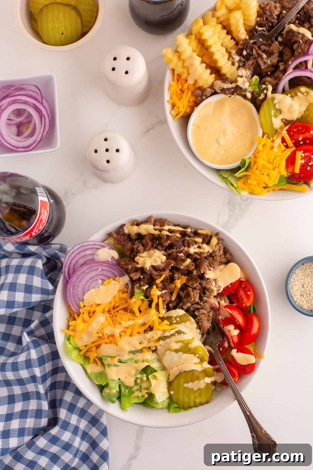Overhead view of two burger bowls filled with cooked ground beef, shredded cheddar cheese, cherry tomatoes, crinkle-cut pickles, sliced red onion, chopped lettuce, and drizzled creamy dressing. Surrounding the bowls are a bottle of Coca-Cola, containers of pickles and onions, salt and pepper shakers, and a blue gingham napkin.