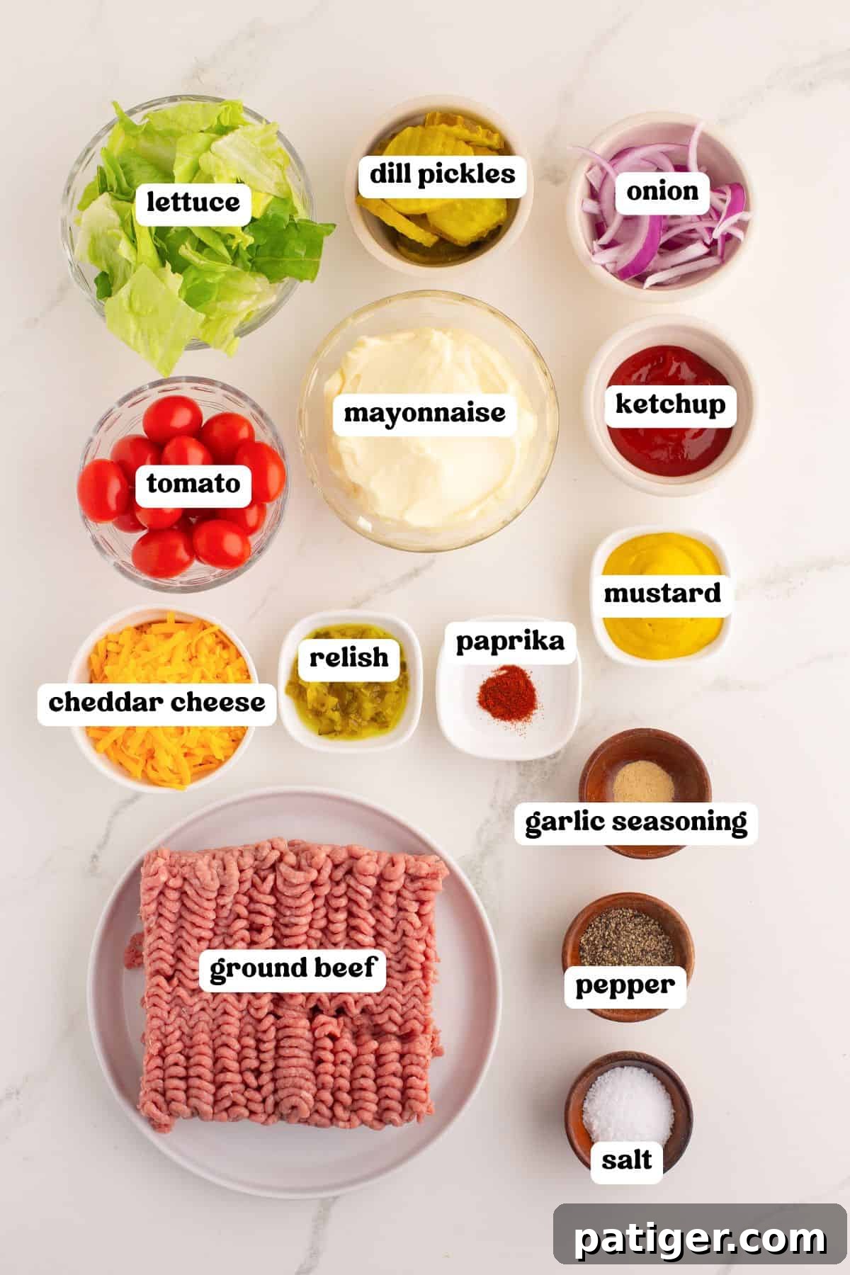 Flat lay of labeled ingredients for burger bowls including: lettuce, dill pickles, onion, tomato, mayonnaise, ketchup, mustard, relish, paprika, garlic seasoning, salt, pepper, cheddar cheese, and raw ground beef.