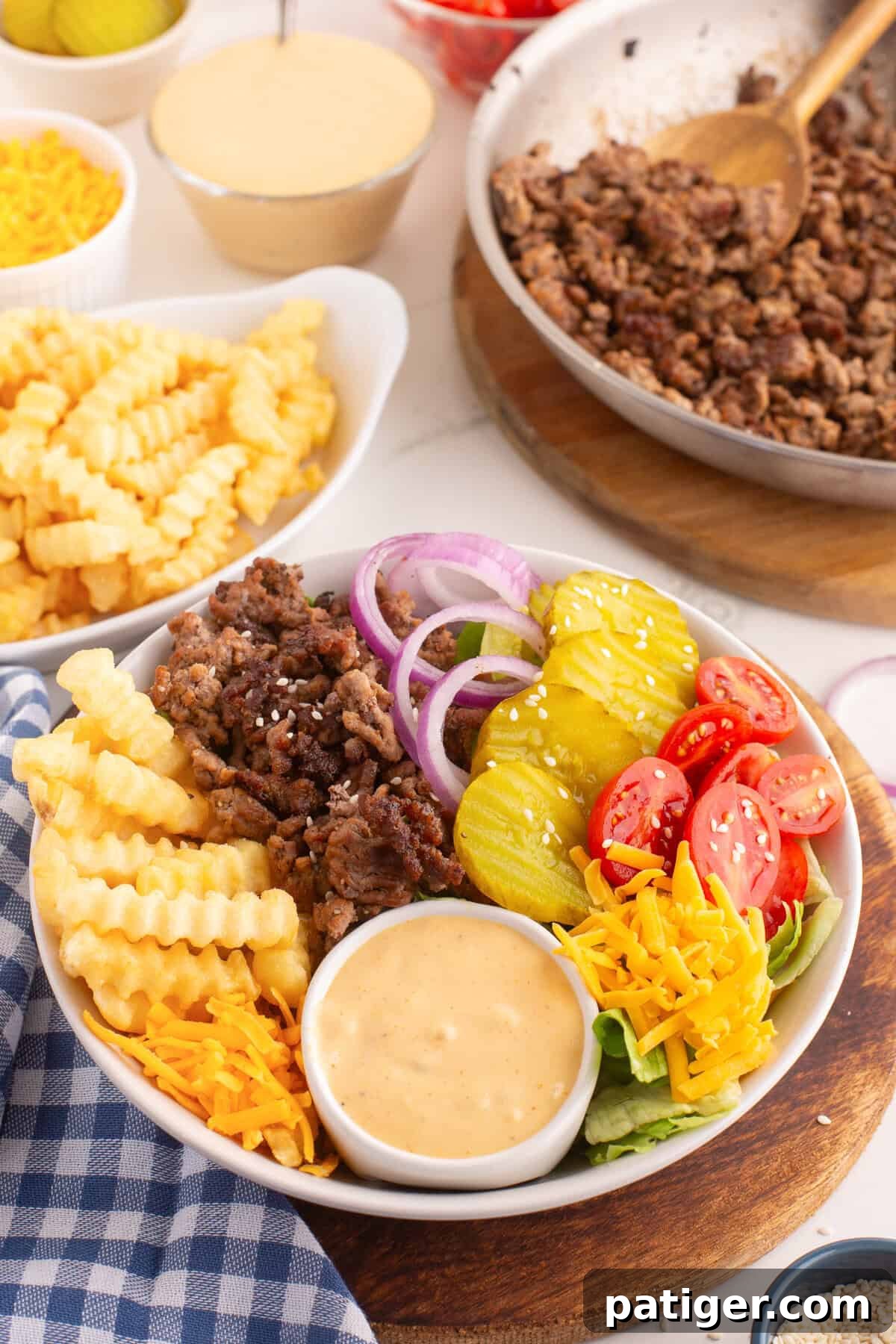 A burger bowl with crinkle-cut French fries, seasoned ground beef, sliced red onions, dill pickles, halved cherry tomatoes, shredded cheddar cheese, lettuce, and a side of creamy special sauce. The bowl is surrounded by dishes of cooked beef, shredded cheese, pickles, tomatoes, and extra fries.
