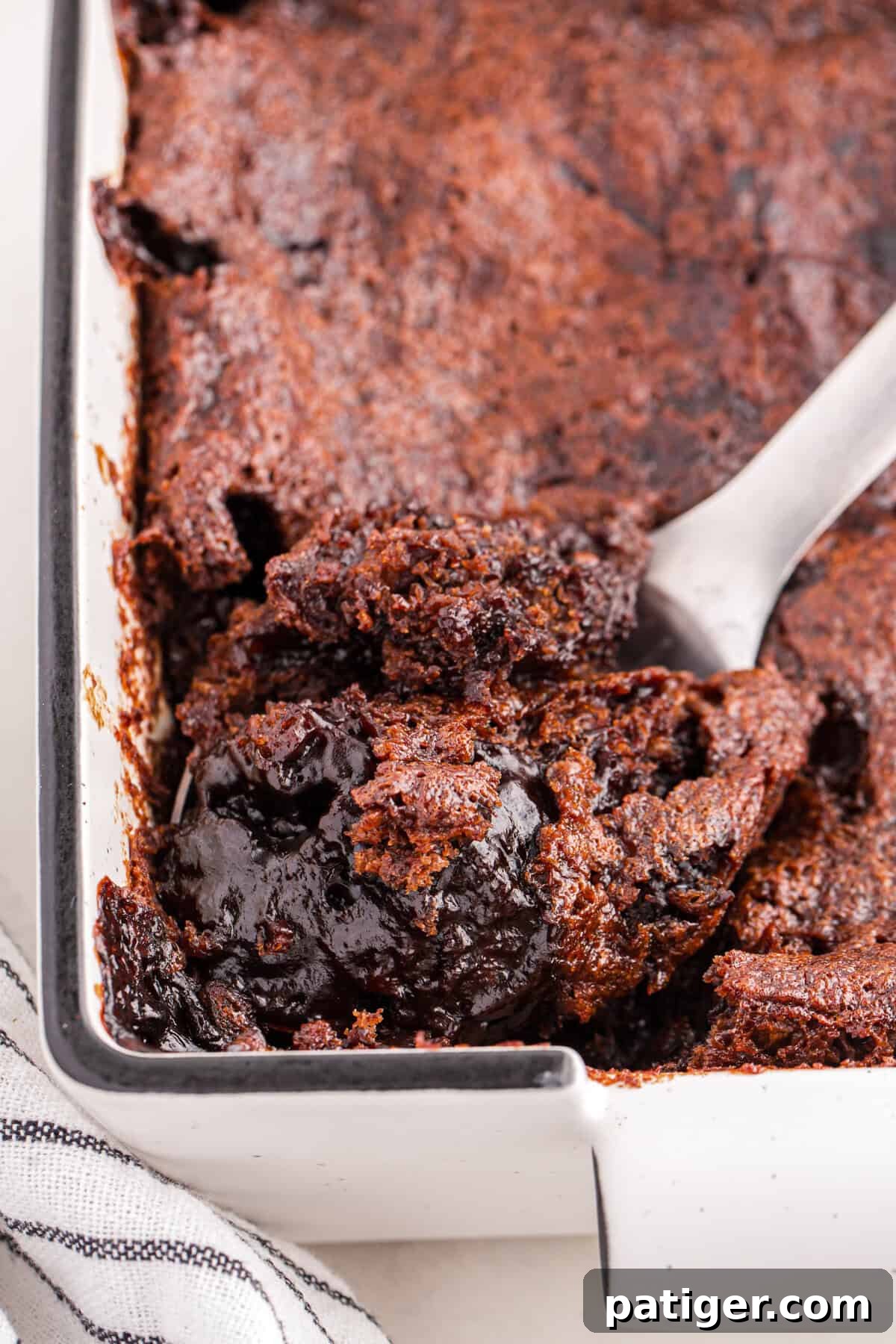 A close-up shot of a rich chocolate pudding cake within a white rectangular baking dish, with a serving spoon poised to scoop out a portion. The image highlights the cake's dark, glossy chocolate sauce layer and its soft, slightly crumbly top texture.