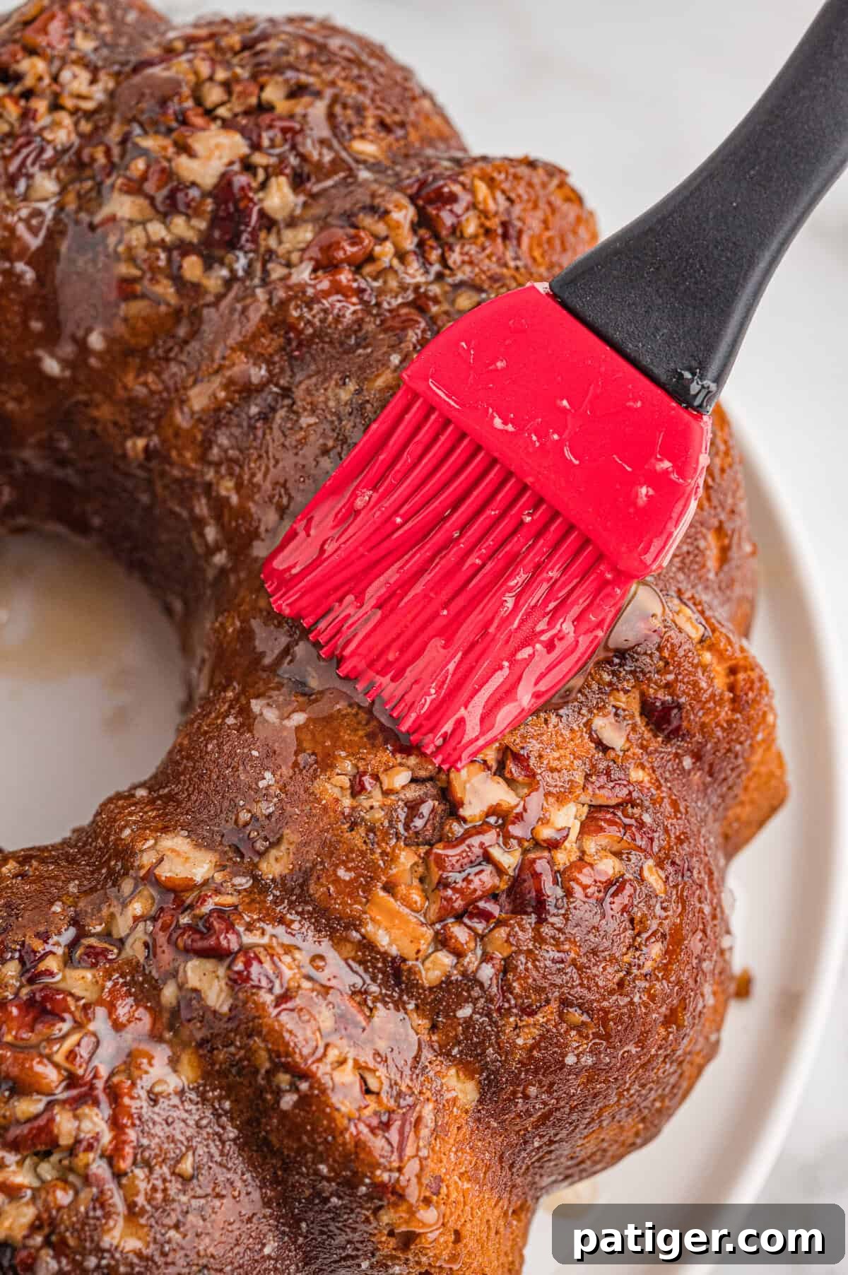Red silicone pastry brush brushing rum glaze onto a baked bundt cake topped with pecans.