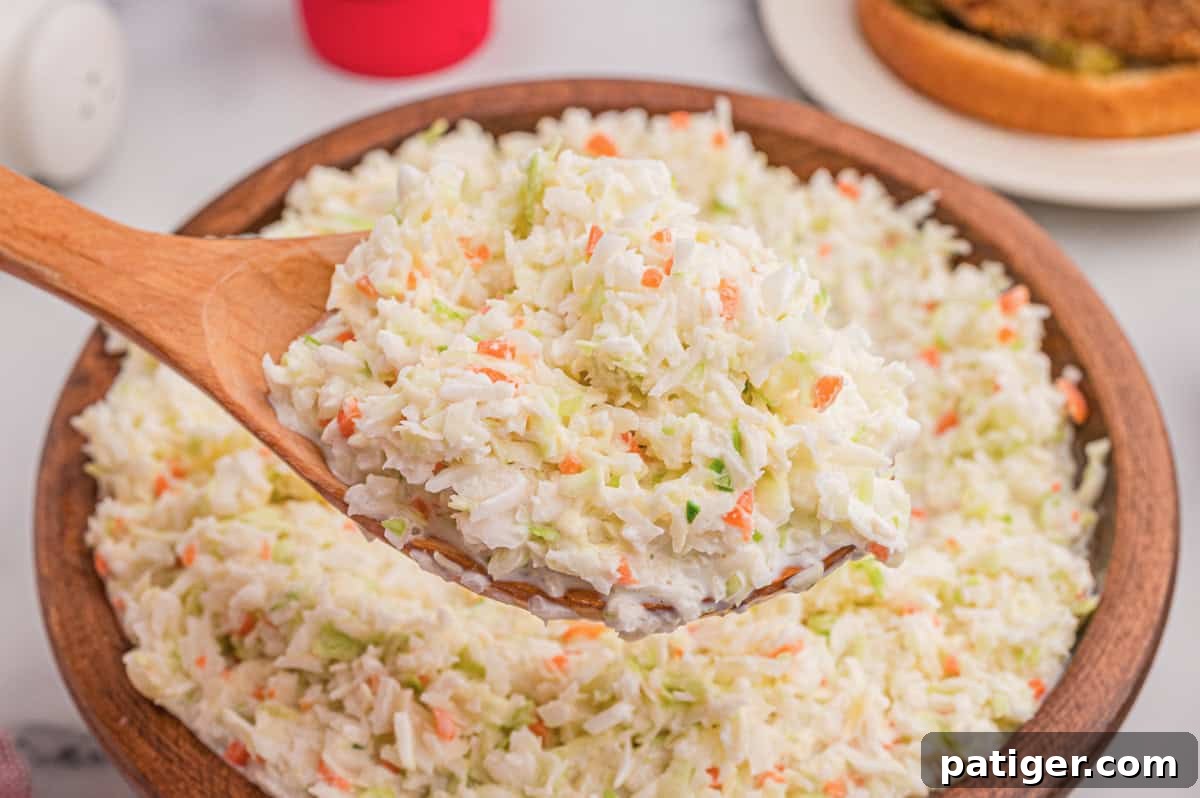 Wooden bowl of coleslaw with a wooden spoon, a chicken sandwich on a plate in the background.