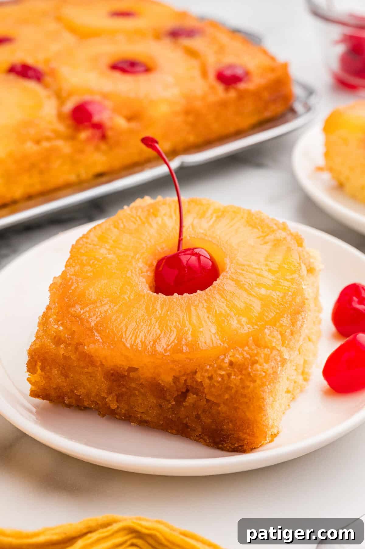 A square slice of pineapple upside down cake on a white plate with a pineapple ring and a maraschino cherry on top.