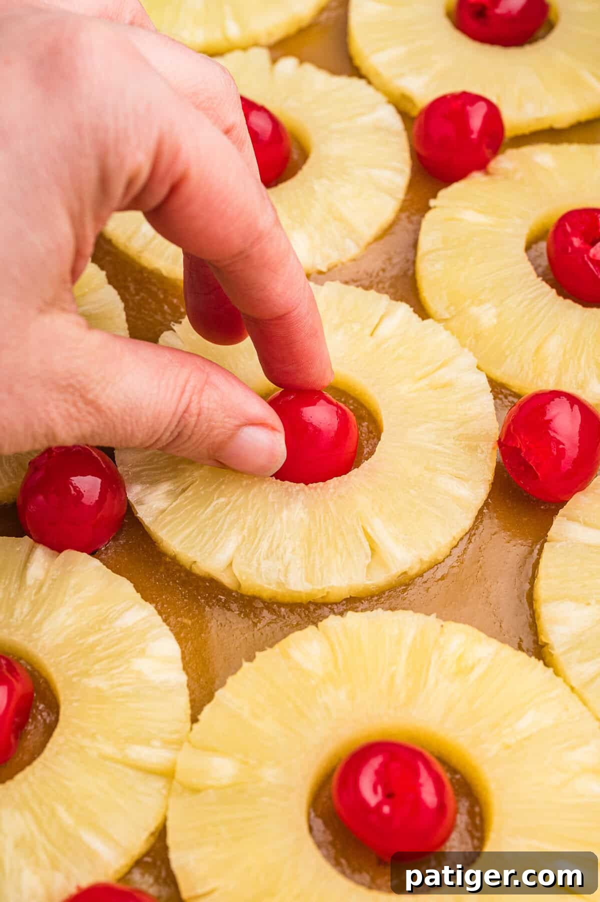 A hand placing a maraschino cherry into the center of a pineapple ring among other arranged rings in a pan.