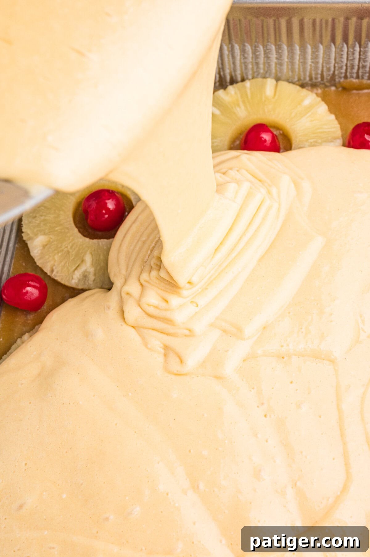 Yellow cake batter being poured over pineapple slices and cherries in a 9x13 baking pan.
