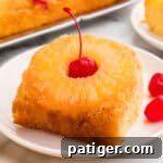 Square plated slice of pineapple upside down cake with a pineapple ring and a cherry on a white dish.