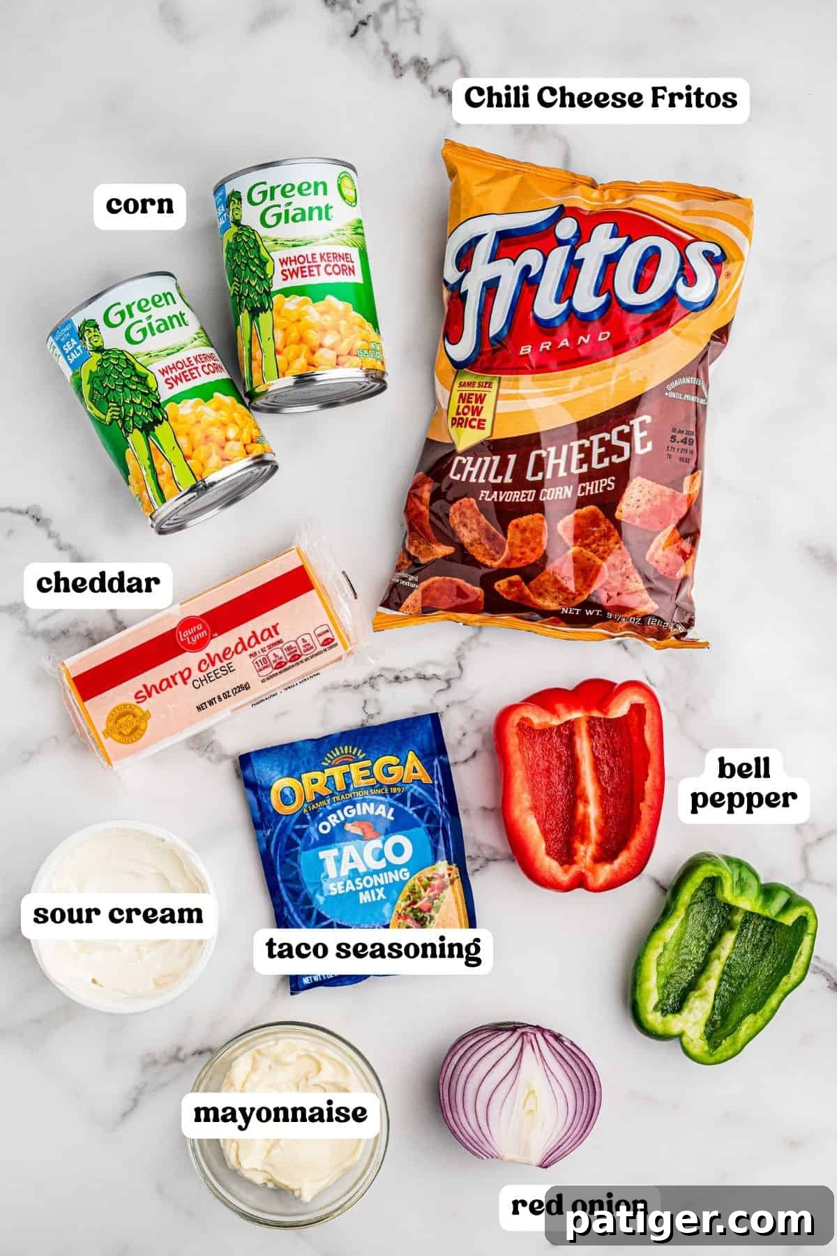 Ingredients on a marble counter: two cans of Green Giant whole kernel corn, a bag of Chili Cheese Fritos, a block of sharp cheddar, sour cream, mayonnaise, an Ortega taco seasoning packet, half a red bell pepper, half a green bell pepper, and half a red onion.
