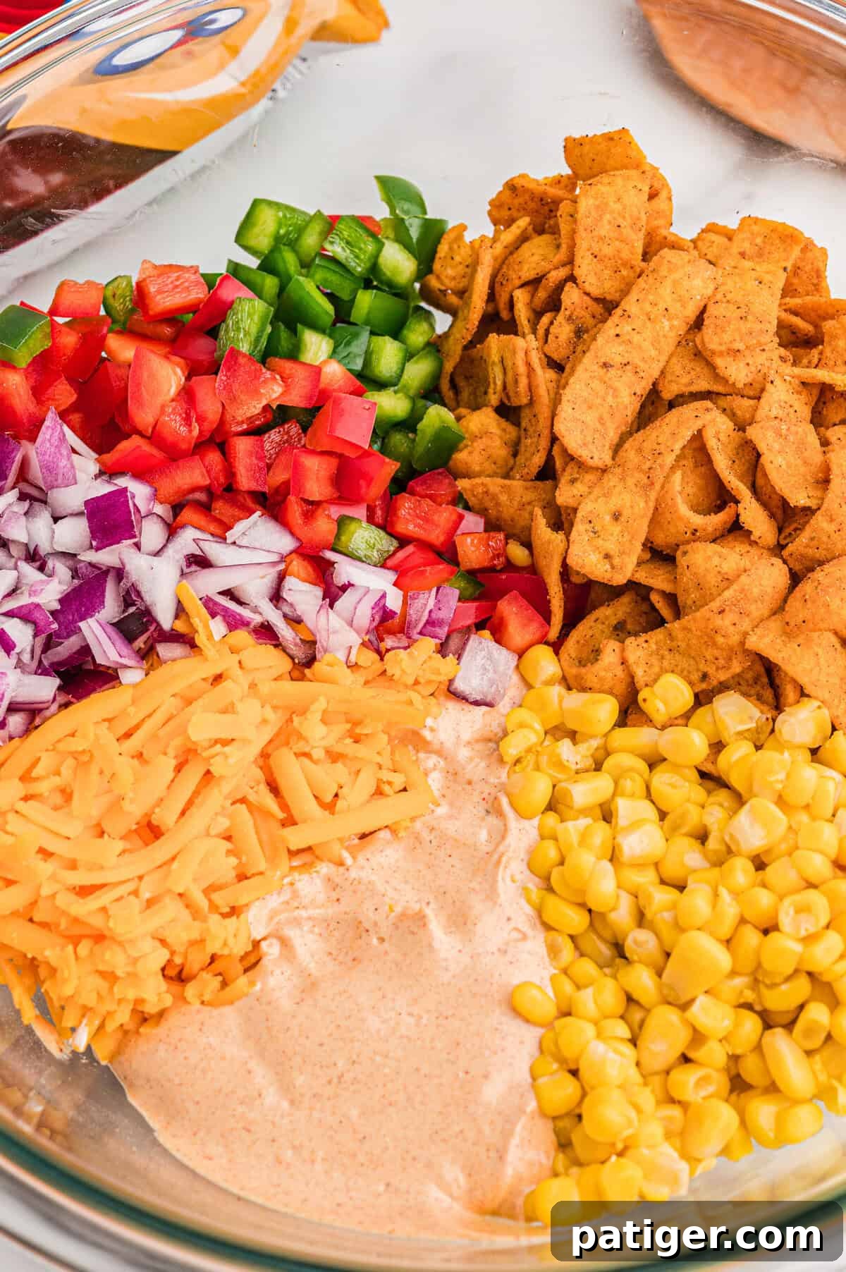 Glass bowl with diced bell pepper, red onion, shredded cheddar, sweet corn, Chili Cheese Fritos, and taco dressing arranged in sections before stirring.