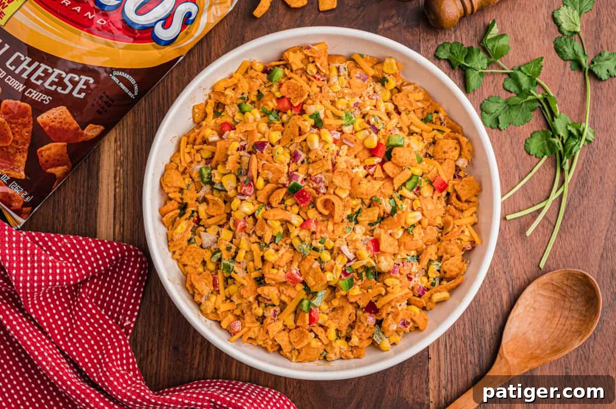 Overhead bowl of Frito salad next to a Chili Cheese Fritos bag, fresh cilantro, a wooden spoon, and a red gingham napkin on a wood board.