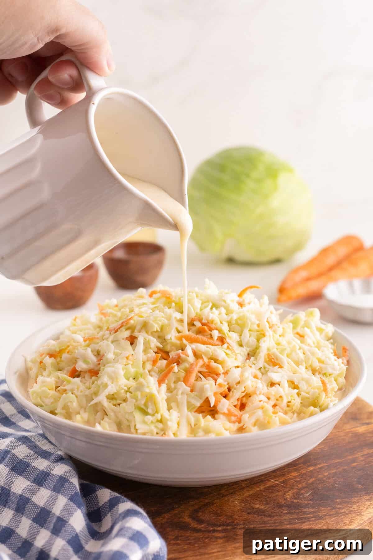 Creamy dressing being poured from a ceramic pitcher onto shredded cabbage and carrots in a white bowl, with a head of cabbage and two carrots in the background.