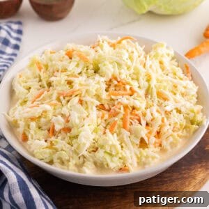 Creamy coleslaw in a white bowl on a wood board with a head of cabbage and carrots in the background.