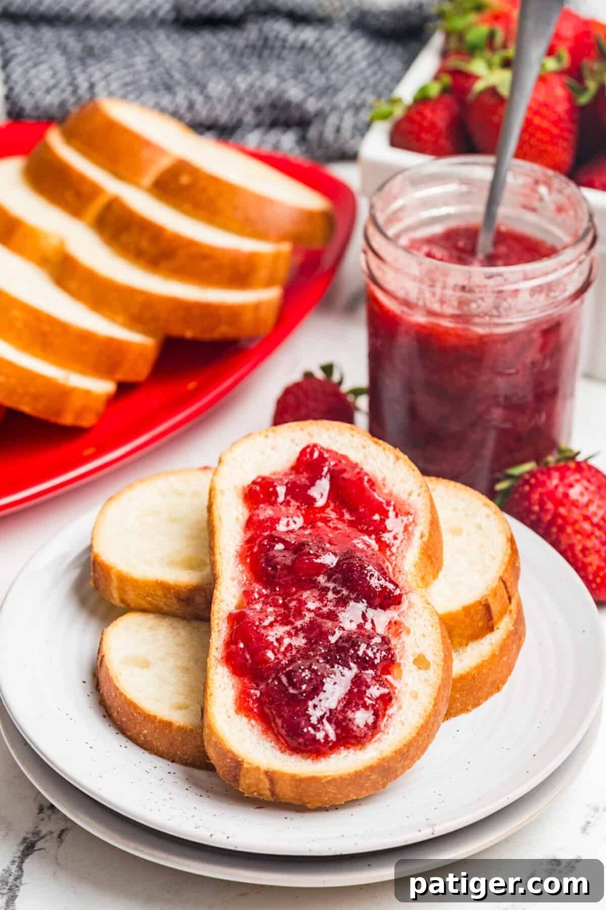 A slice of bread topped with strawberry jam in front of a red platter of bread slices, a jar of jam with a spoon, and fresh strawberries.
