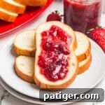 Bread slices topped with homemade strawberry jam stacked on a white plate with a jar of jam nearby.