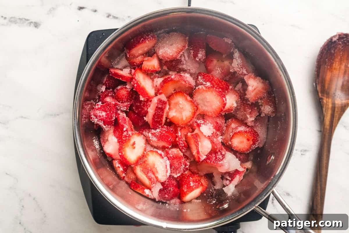 Sliced strawberries in a saucepan sprinkled with granulated sugar with a wooden spoon to the side.