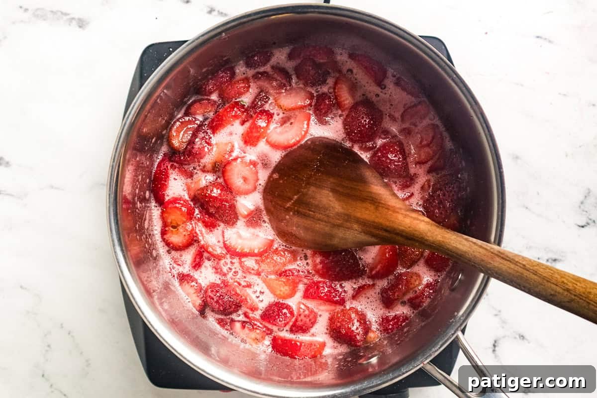 Softened sliced strawberries simmering in a saucepan with a wooden spoon stirring the bright pink mixture.