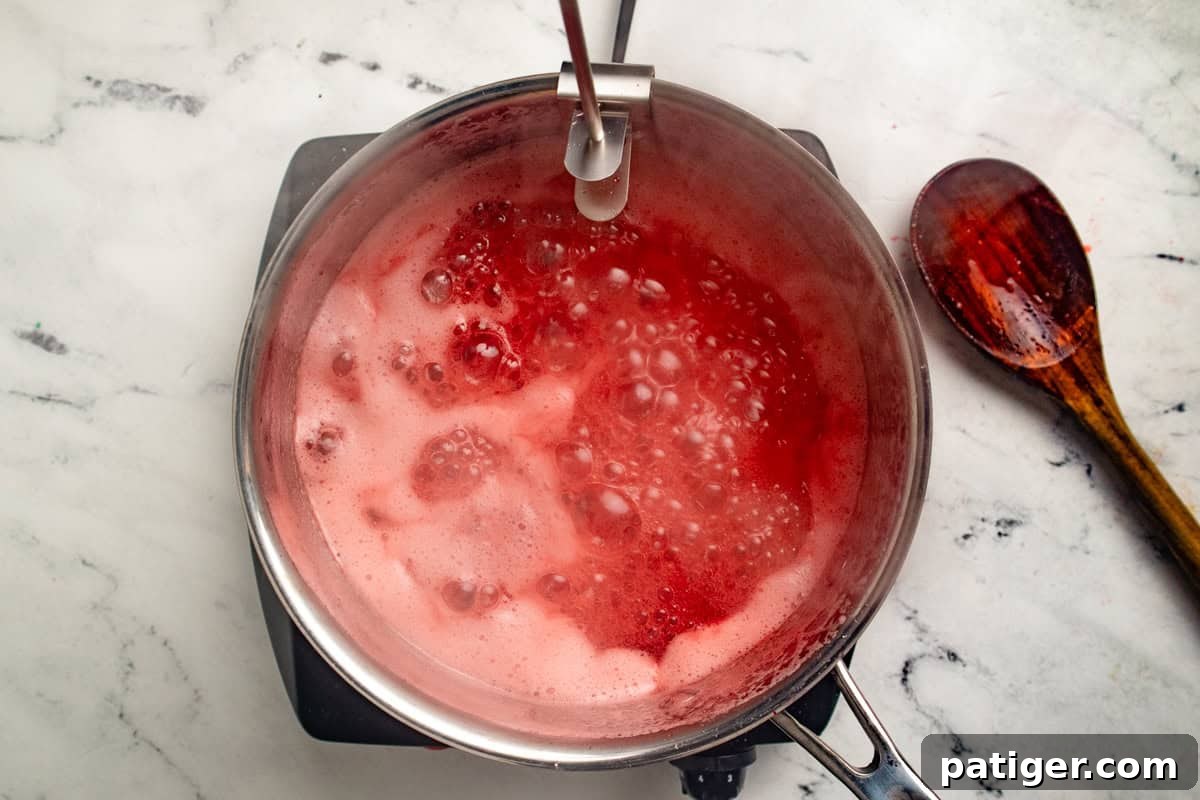 Bubbling strawberry jam in a saucepan with a candy thermometer attached to the rim.