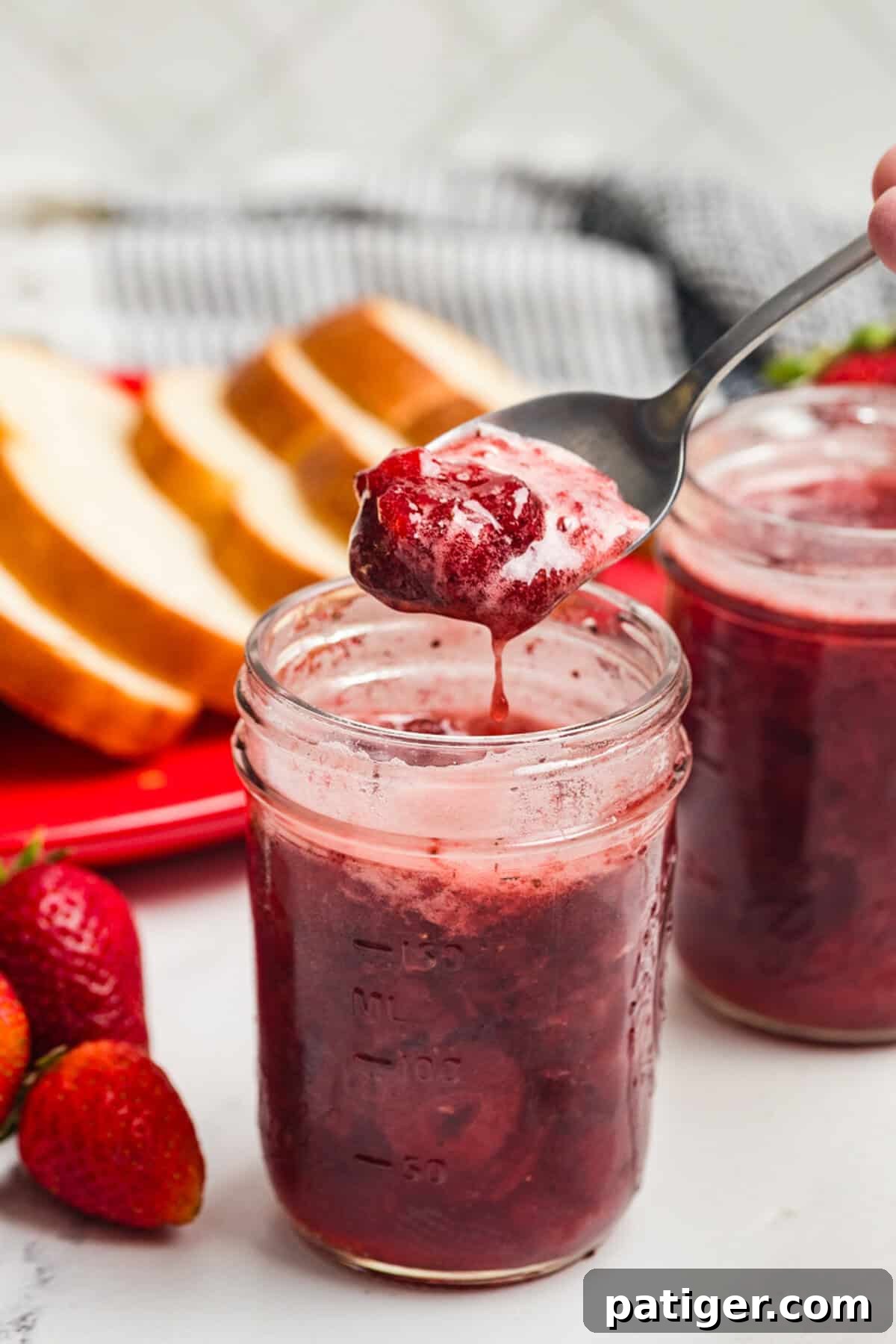 A spoon lifting strawberry jam from an open mason jar with bread slices nearby.