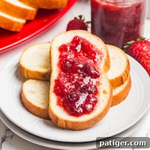 Bread slices topped with homemade strawberry jam stacked on a white plate with a jar of jam nearby.