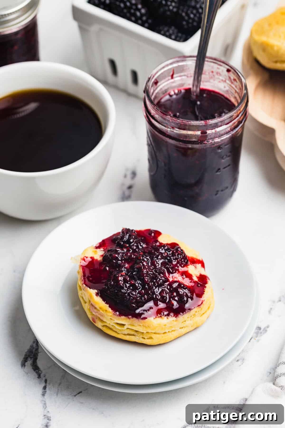 A split biscuit topped with blackberry jam on a white plate with a jar of jam and a cup of coffee in the background.