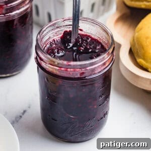 A close-up square shot of an open mason jar of blackberry jam with a spoon stirring the surface.