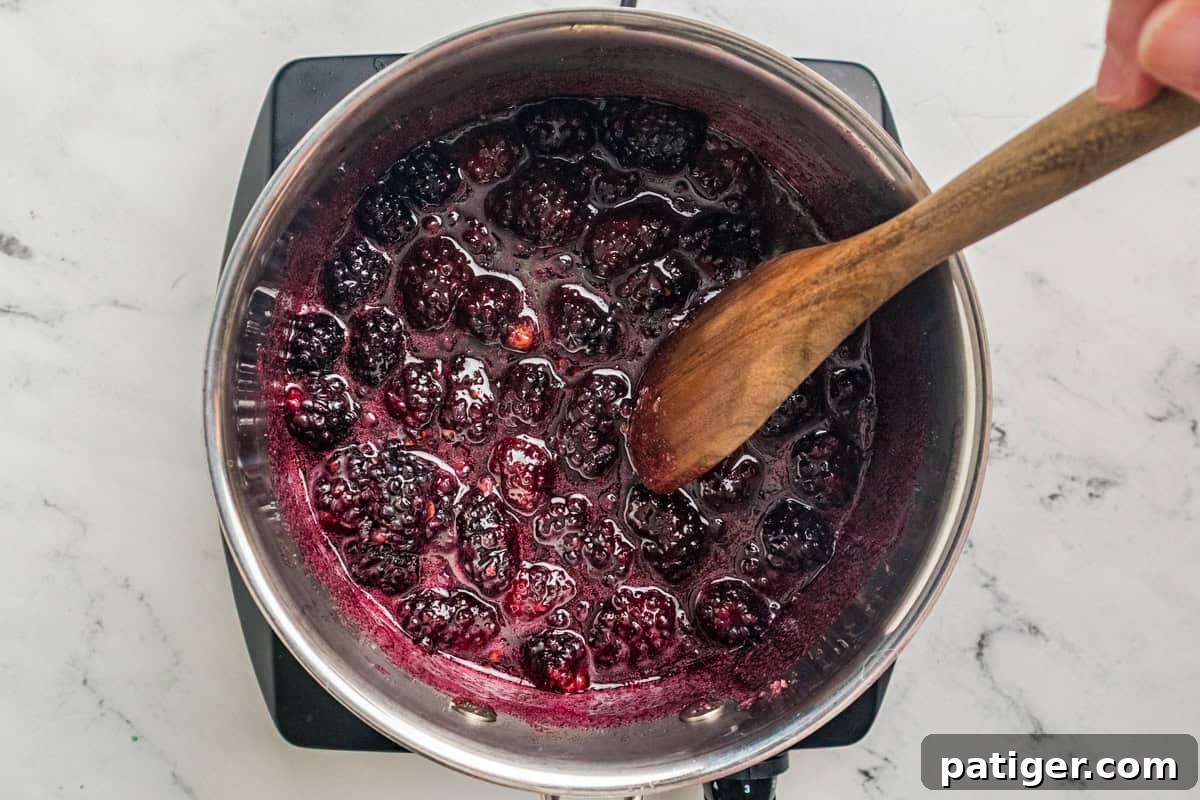 Whole blackberries breaking down in a saucepan with a wooden spoon stirring the dark juice.