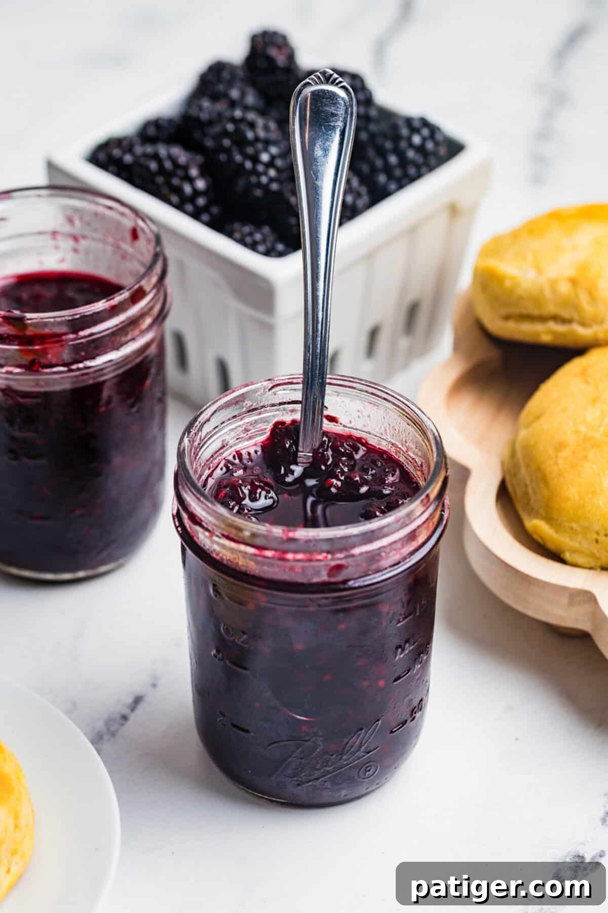 A tall open mason jar of blackberry jam with a spoon resting in it and a basket of fresh blackberries in the background.