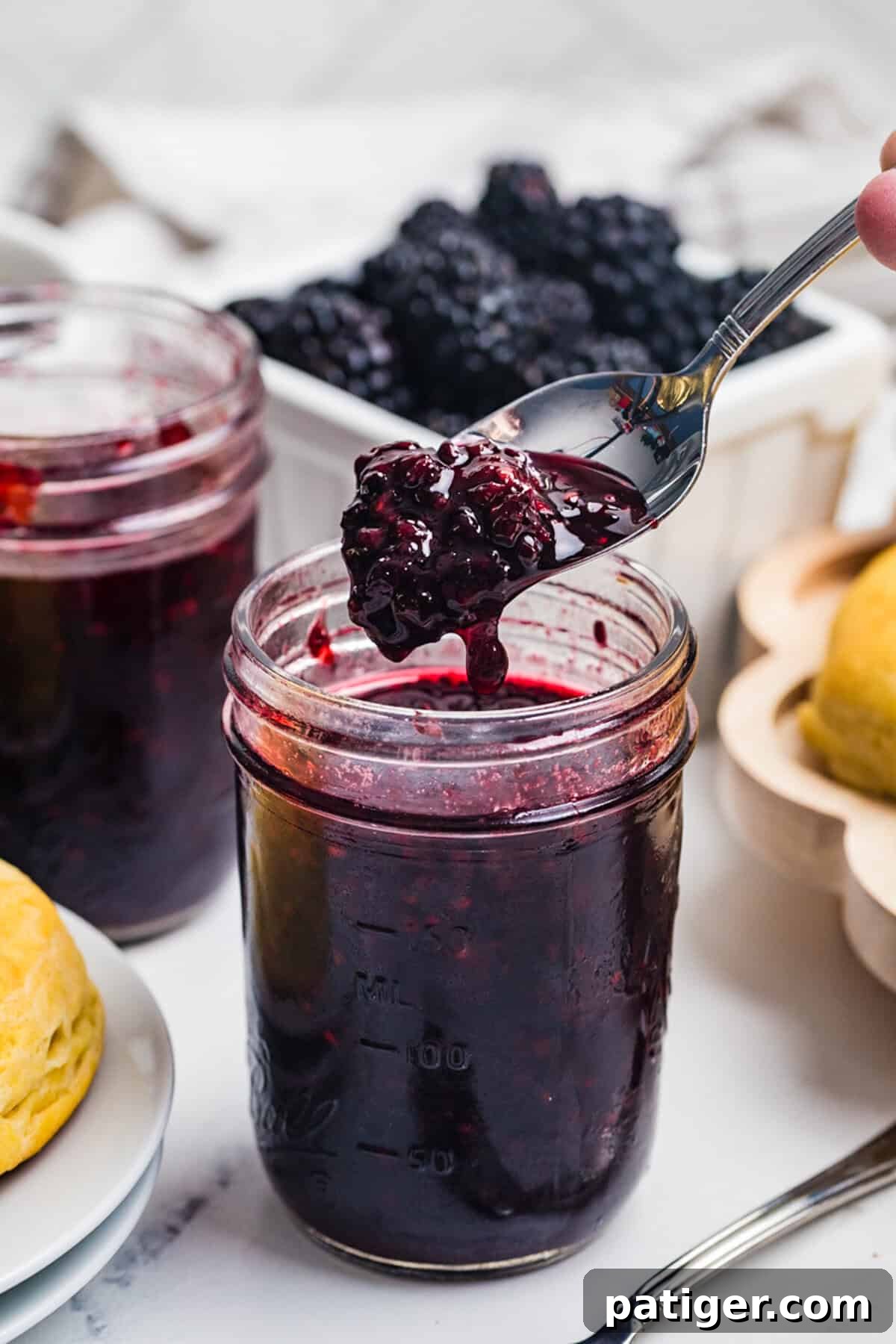 A spoon lifting a thick scoop of blackberry jam from a mason jar with a basket of fresh blackberries behind it.