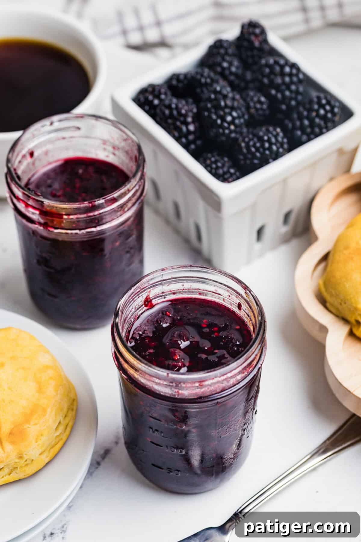 Two open jars of homemade blackberry jam on a counter with a basket of fresh blackberries, a coffee cup, and a biscuit.