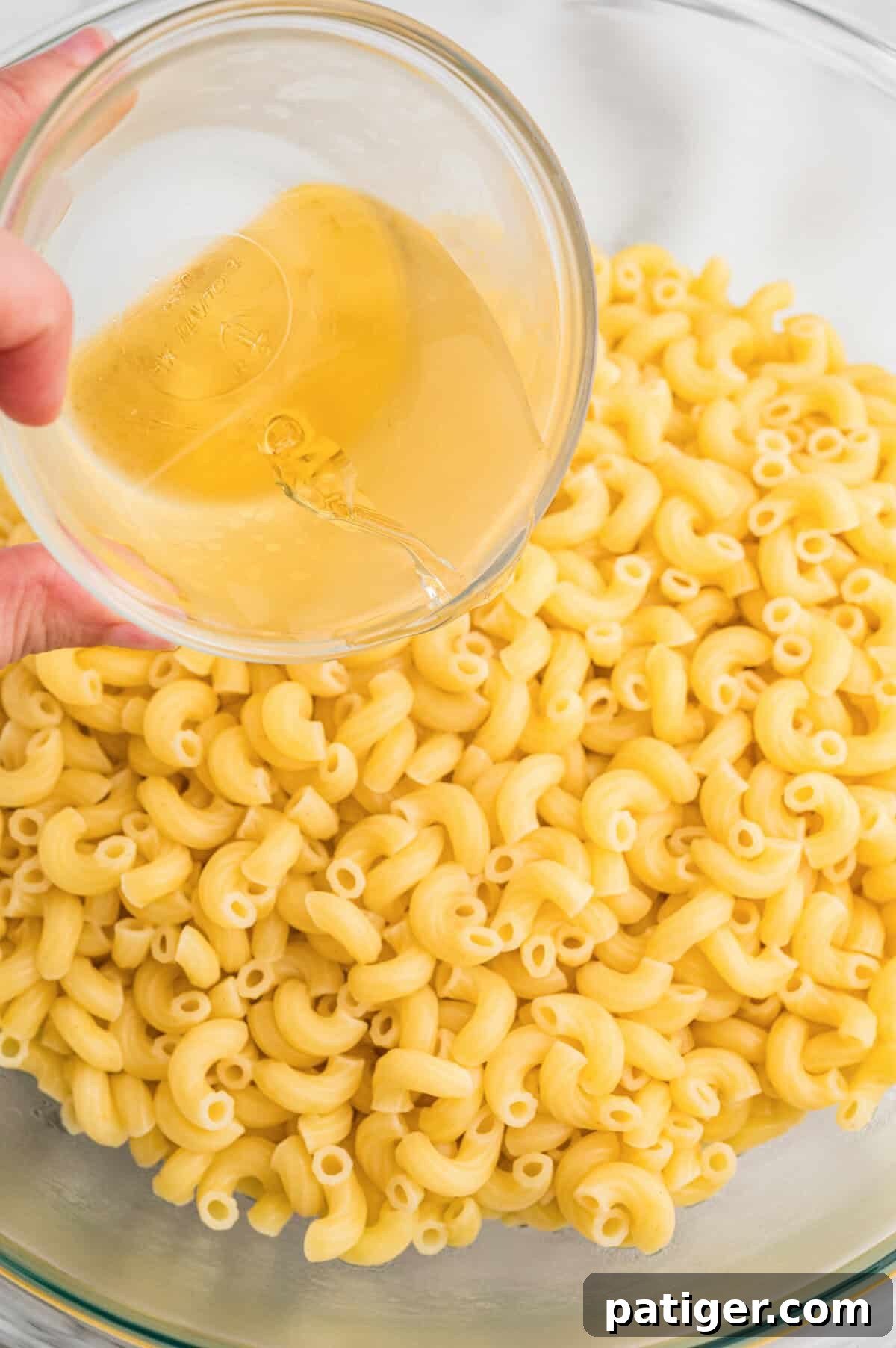Apple cider vinegar being poured from a small glass bowl over cooked elbow macaroni in a large glass mixing bowl.