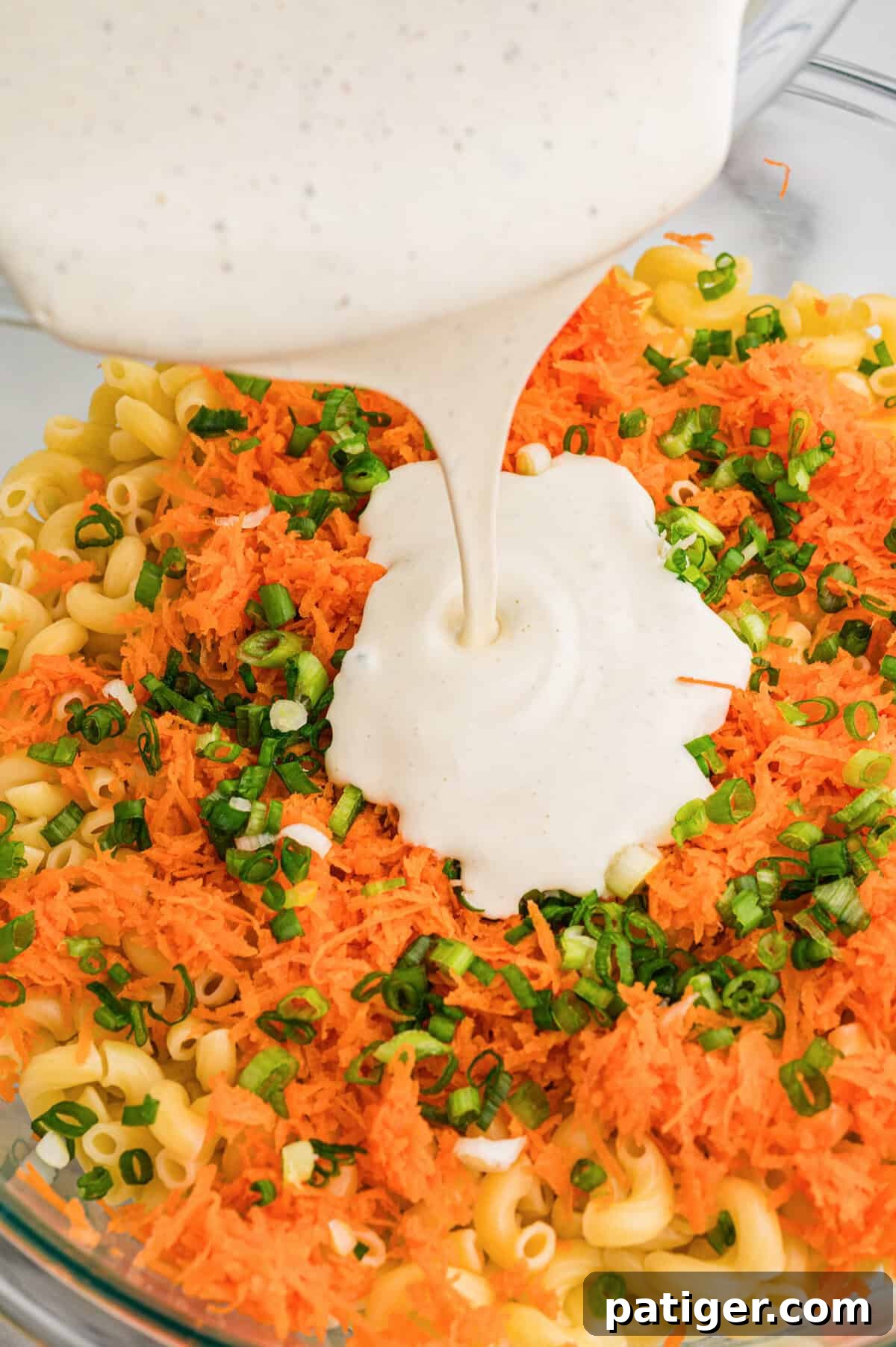 Creamy dressing being poured over cooked macaroni, shredded carrots, and green onions in a large glass bowl.