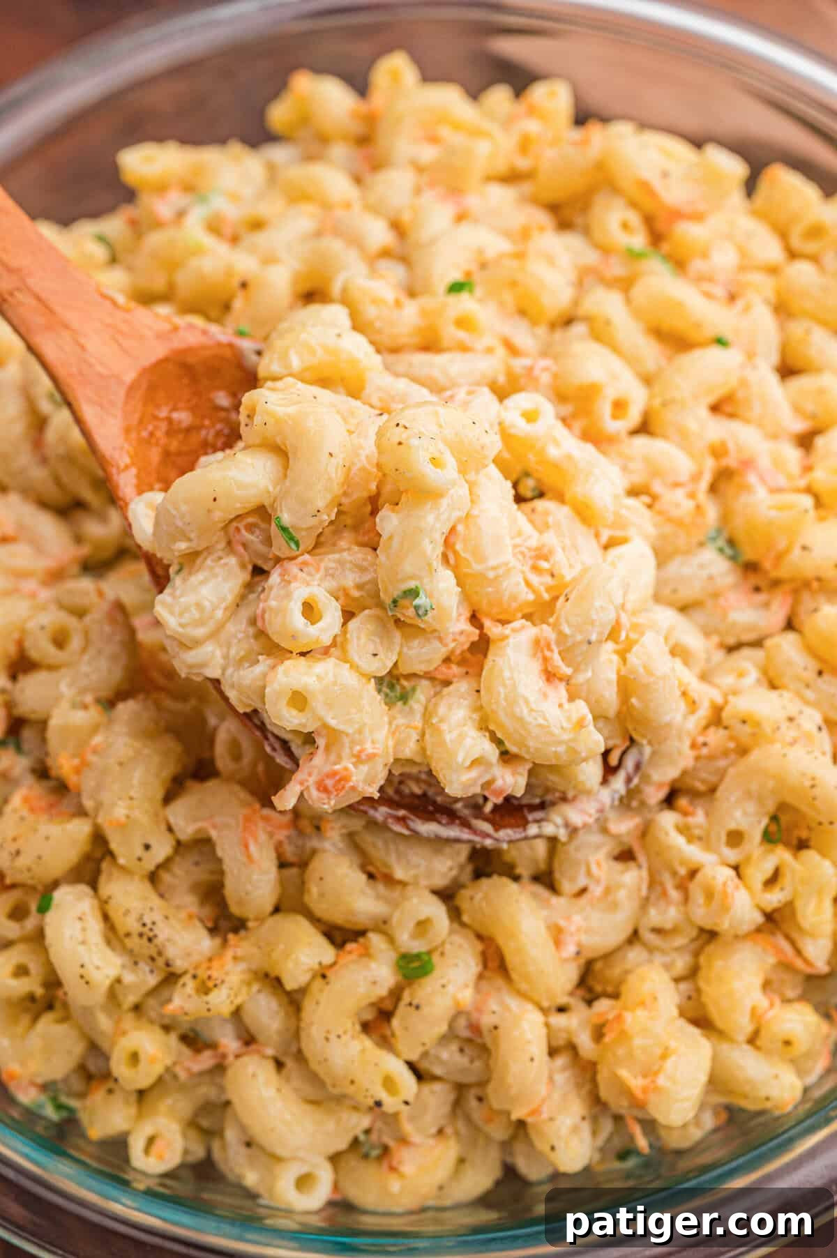 A wooden spoon lifting a scoop of creamy mac salad with shredded carrots and green onion from a clear glass bowl.