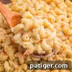 A wooden spoon lifting a scoop of creamy Hawaiian mac salad with shredded carrots and green onion from a bowl.