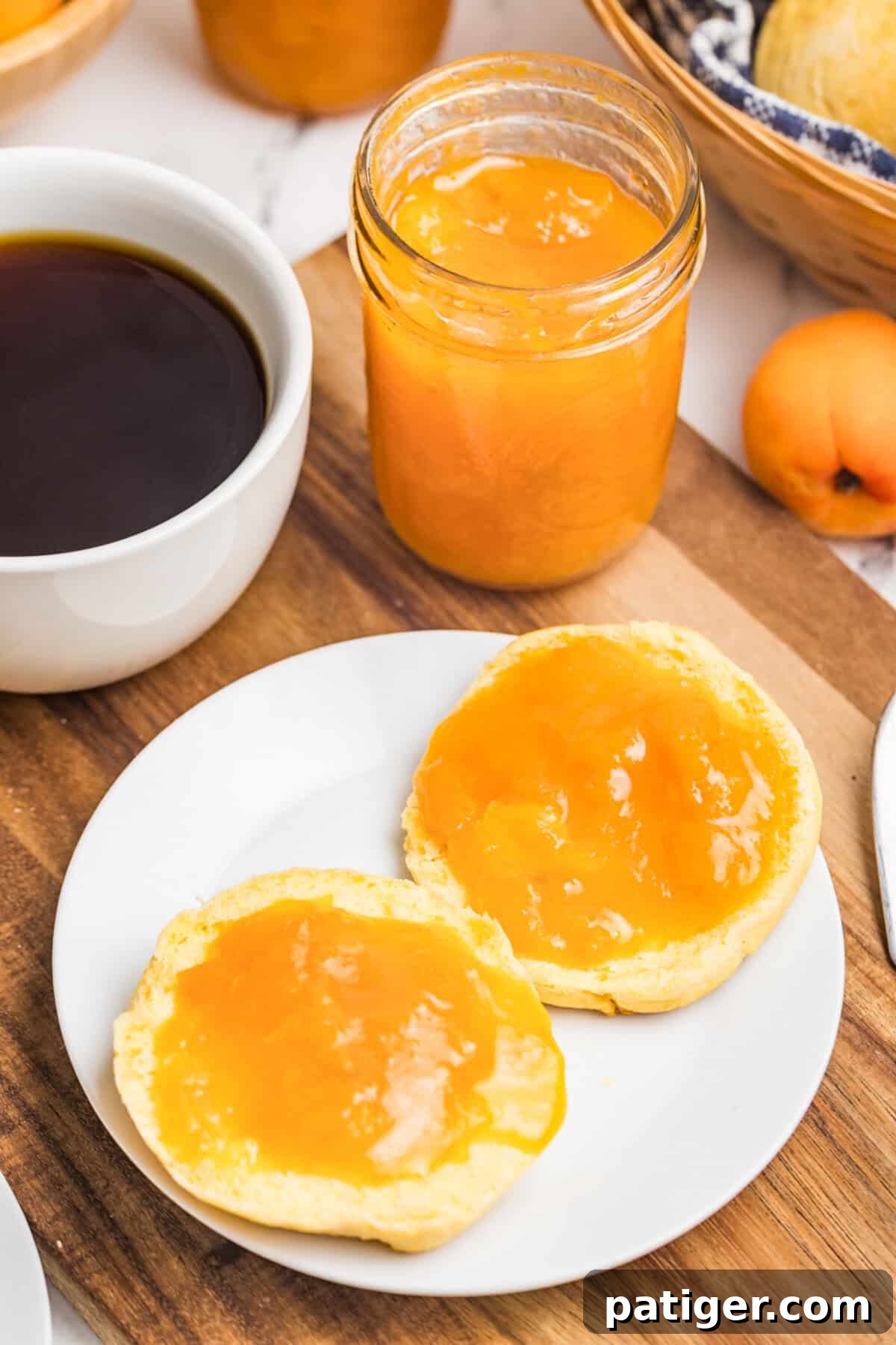 Apricot Jam Without Pectin: Simple Homemade Recipe 3 An open mason jar of apricot jam on a wooden board with two biscuit halves spread with jam on a white plate, a cup of black coffee, and a fresh apricot beside it.