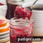 Homemade Raspberry Jam Without Pectin — Simple Stove-Top Recipe 10 A close-up square shot of a spoon lifting raspberry jam from a small mason jar with a slice of bread in the background.
