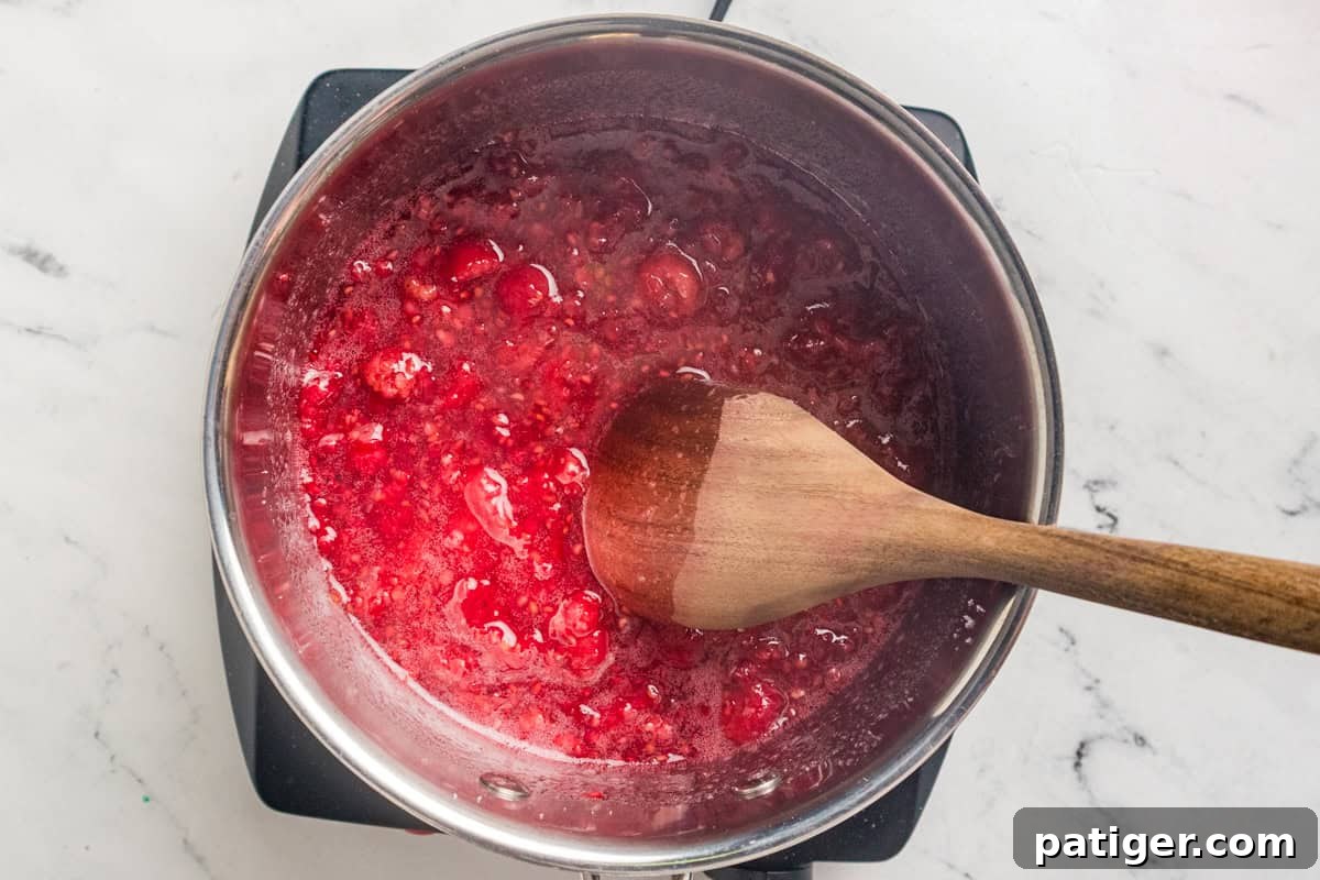 Homemade Raspberry Jam Without Pectin — Simple Stove-Top Recipe 4 Raspberries breaking down in a saucepan with a wooden spoon stirring the bright red juice.