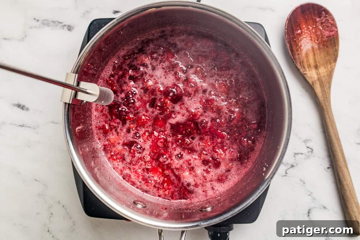 Homemade Raspberry Jam Without Pectin — Simple Stove-Top Recipe 5 Bubbling raspberry jam in a saucepan with a candy thermometer attached and a wooden spoon to the side.