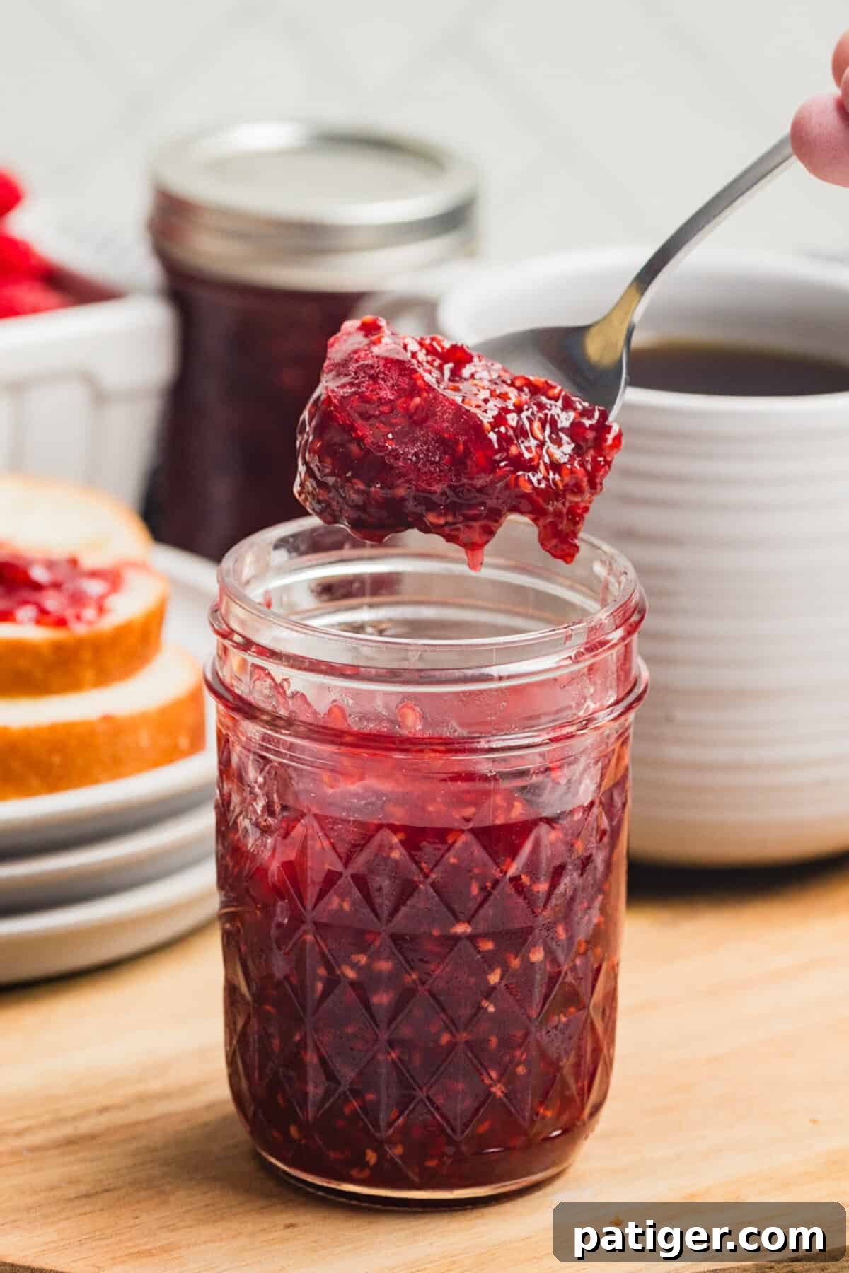 Homemade Raspberry Jam Without Pectin — Simple Stove-Top Recipe 7 A spoon lifting raspberry jam from a small mason jar with a slice of bread and jar of jam in the background and a coffee mug to the side.