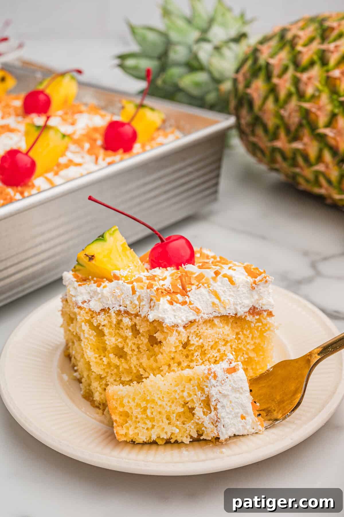 Tropical Piña Colada Layer Cake with Rum-Soaked Pineapple 2 A slice of pina colada cake on a plate topped with whipped topping, toasted coconut, a pineapple wedge, and a cherry with a fresh pineapple in the background.