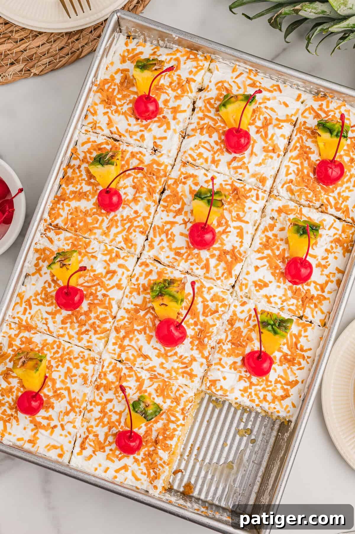 Tropical Piña Colada Layer Cake with Rum-Soaked Pineapple 3 Top-down view of the cake in a pan with one corner piece removed, squares topped with toasted coconut, cherries, and pineapple.