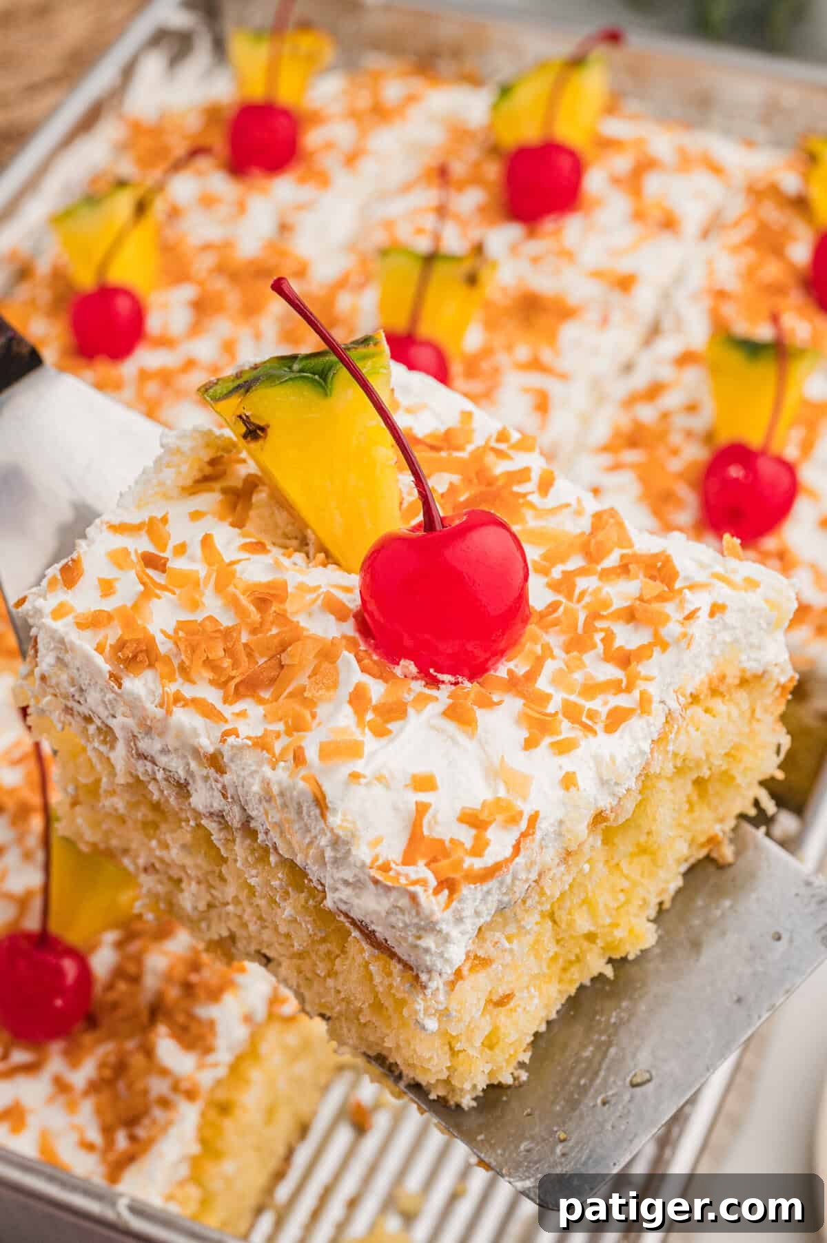 Tropical Piña Colada Layer Cake with Rum-Soaked Pineapple 8 A slice of poke cake lifted on a metal server, showing yellow cake, whipped topping, toasted coconut, and a cherry.