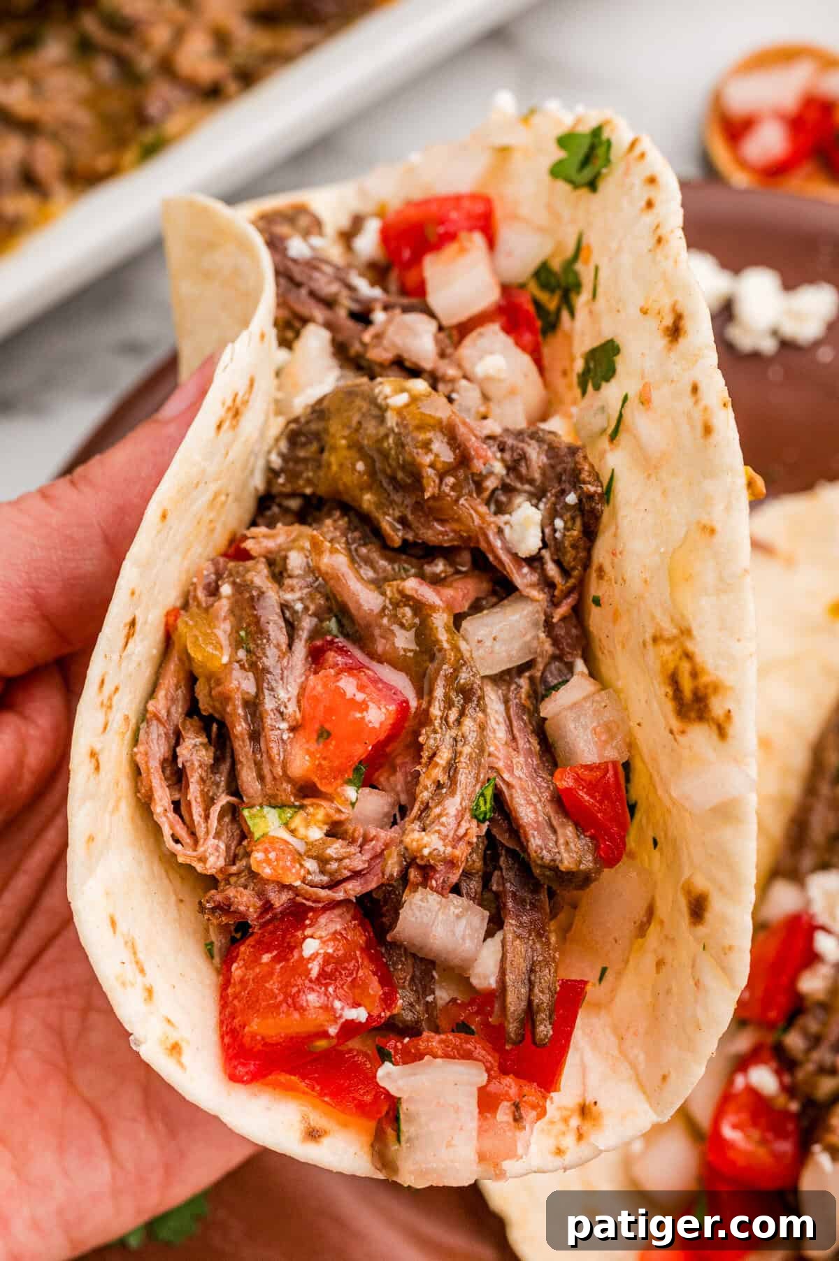 Juicy Slow Cooker Carne Asada Recipe 11 A hand holding a soft taco filled with shredded carne asada, diced tomato, onion, and crumbled cotija cheese.